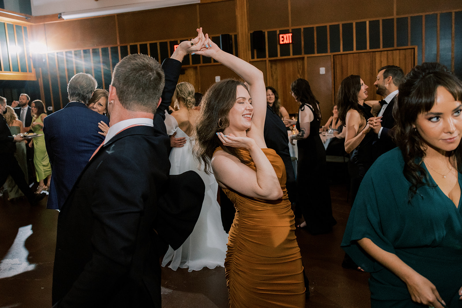 People dancing at an indoor event, with a woman in a brown dress striking a playful pose in the center of the image.