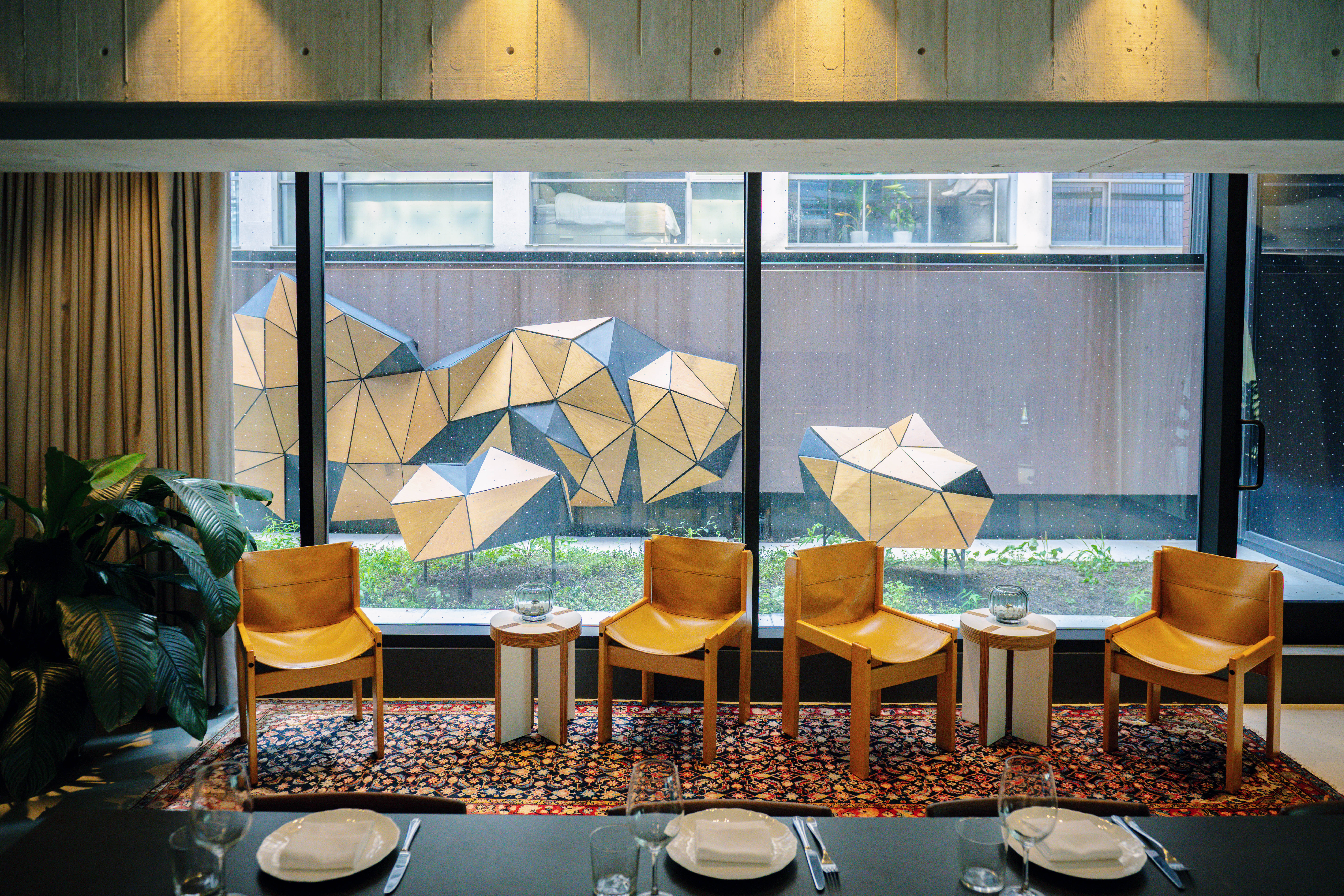 A modern dining room with a set table, wooden chairs, and large windows overlooking a courtyard with geometric sculptures.