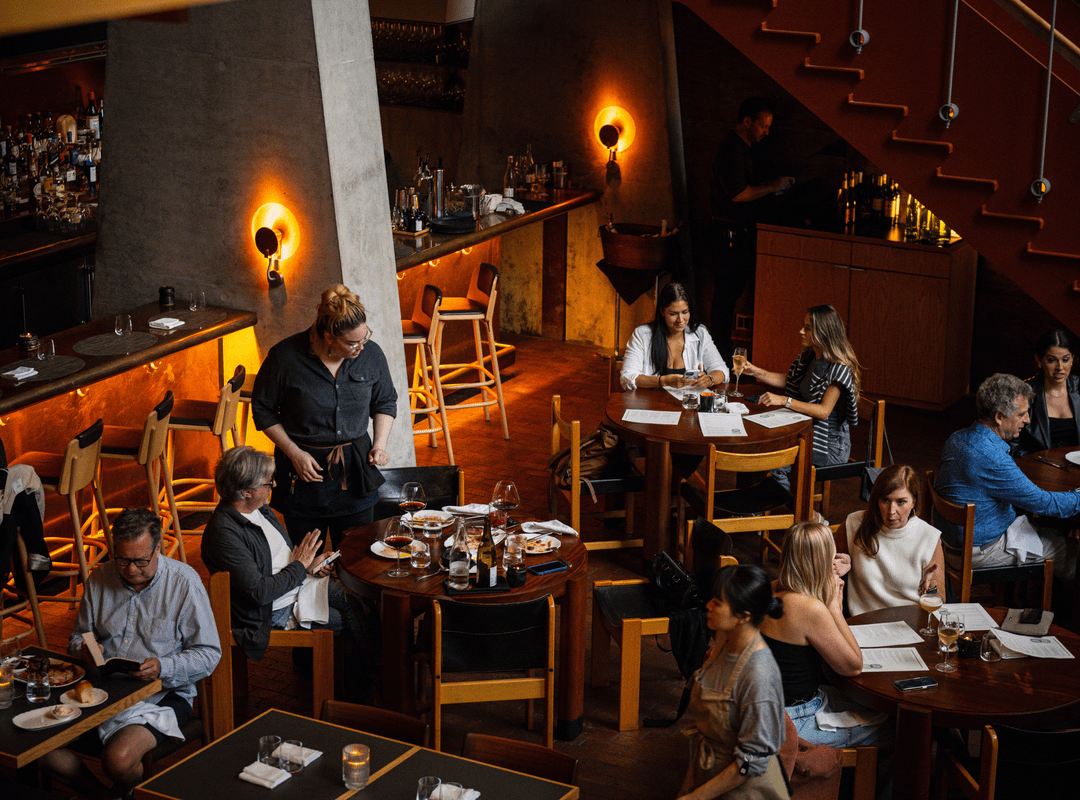 A busy restaurant with diners seated at tables, a server attending guests, and warm ambient lighting under a staircase.