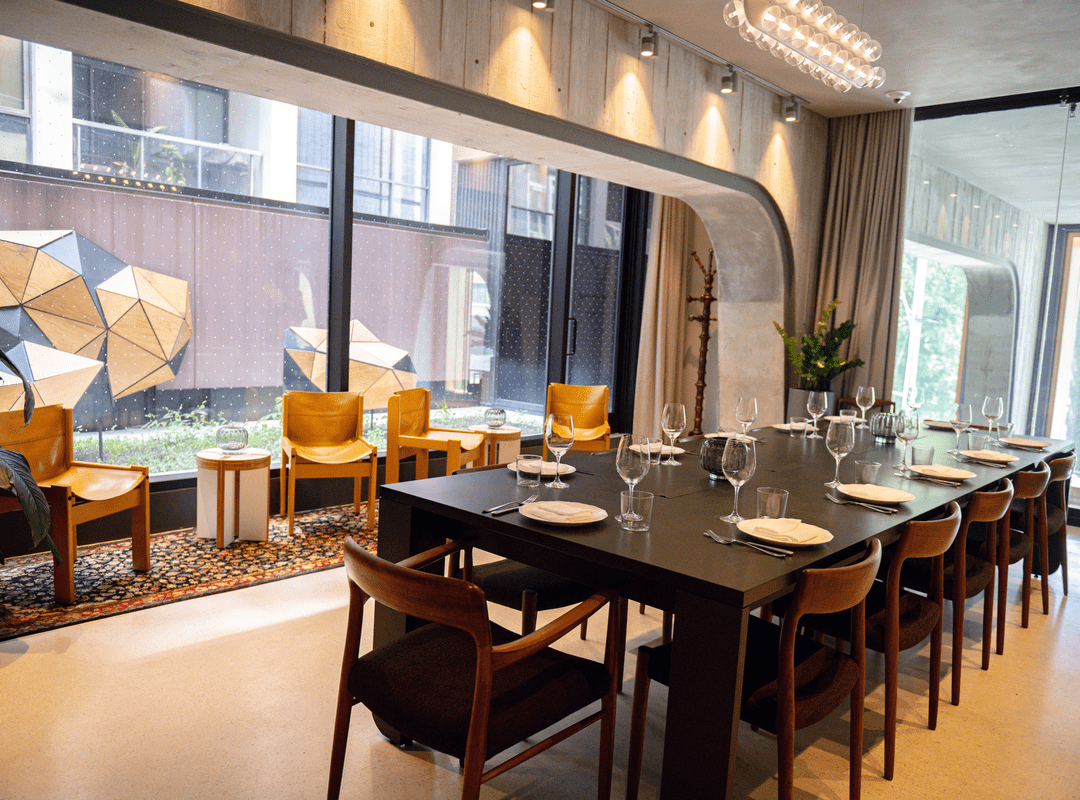 A modern dining room with a set table, wooden chairs, and large windows overlooking a courtyard with geometric sculptures.