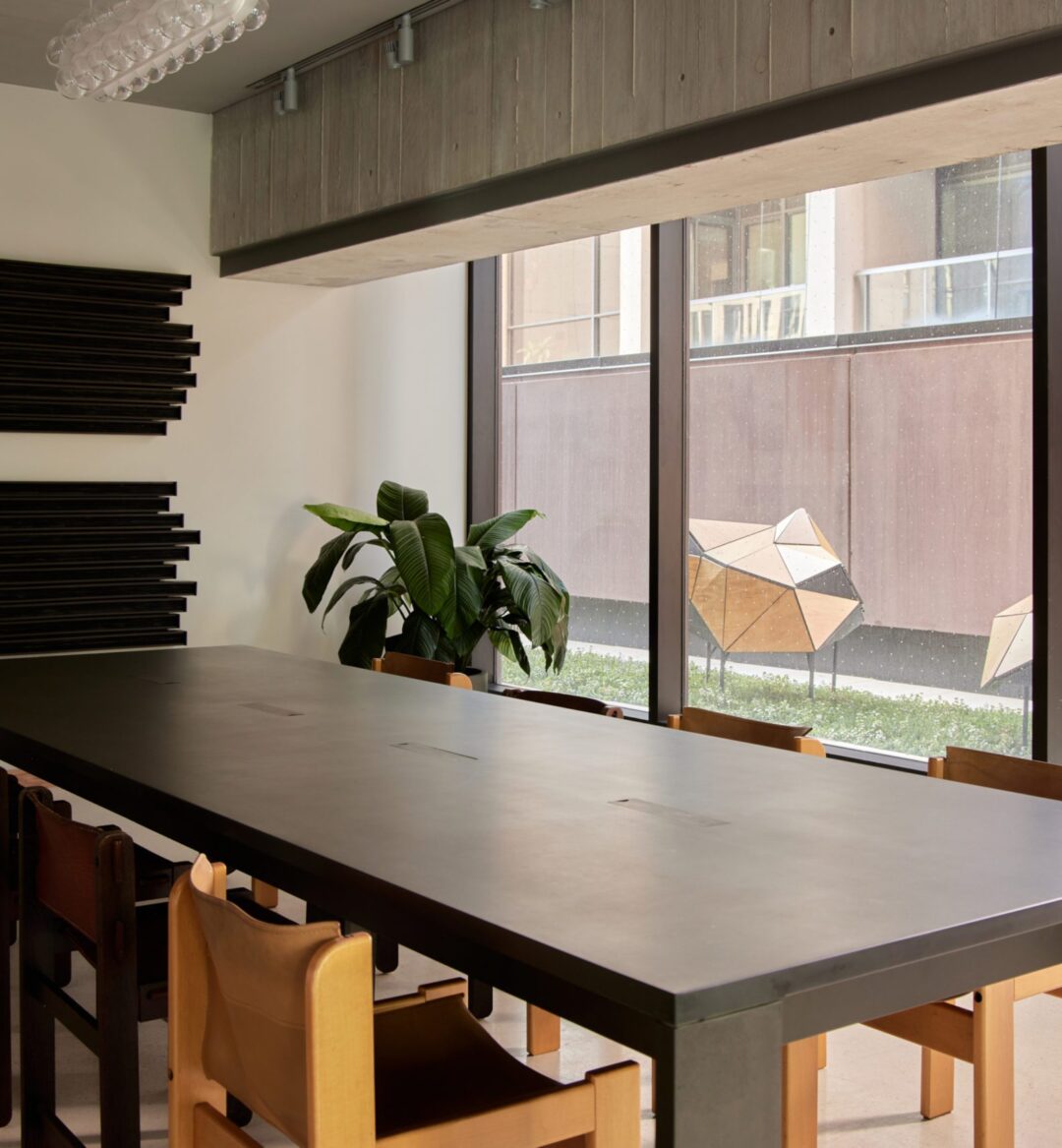 A modern conference room with a long black table, wooden chairs, a plant, large windows, and geometric wall art.