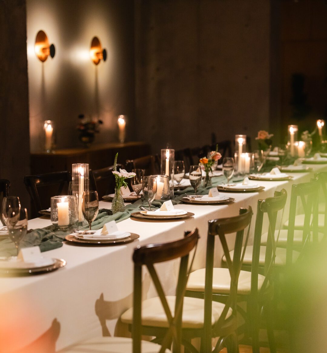 A long dining table set with plates, glasses, candles, small flower arrangements, and place cards in a dimly lit room.
